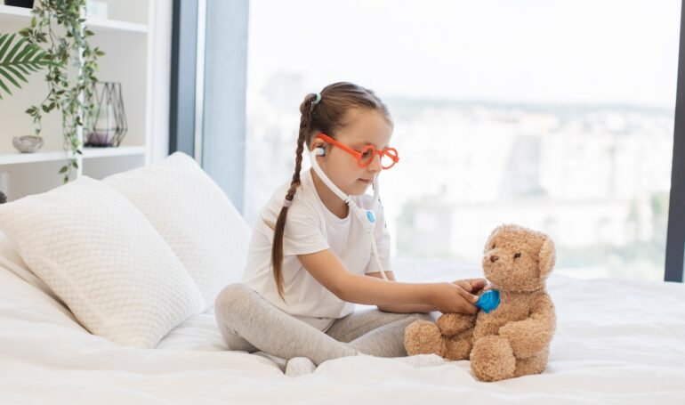 Girl with toy doctor kit examining teddy on bed in own room