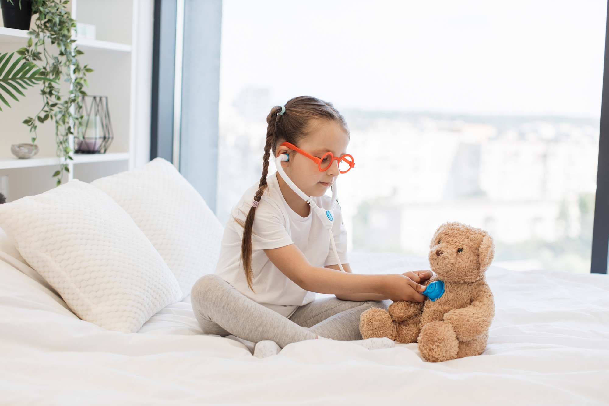 Girl with toy doctor kit examining teddy on bed in own room