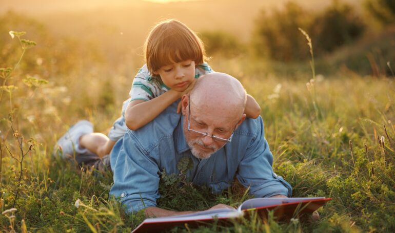 Little boy with his grandfather reading a book in a field at summer